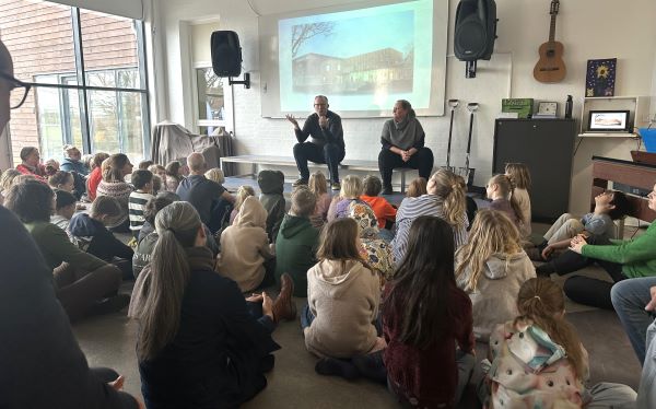 To voksne sidder på en scene og fortæller, mens en masse børn og enkelte voksne kigger på.